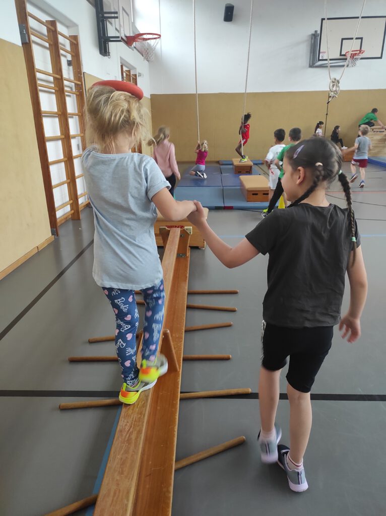 Kinder turnen in der Turnhalle an Stationen, ein Kindergartenkind balanciert auf Langbank, ein Schulkind gibt Hilfestellung