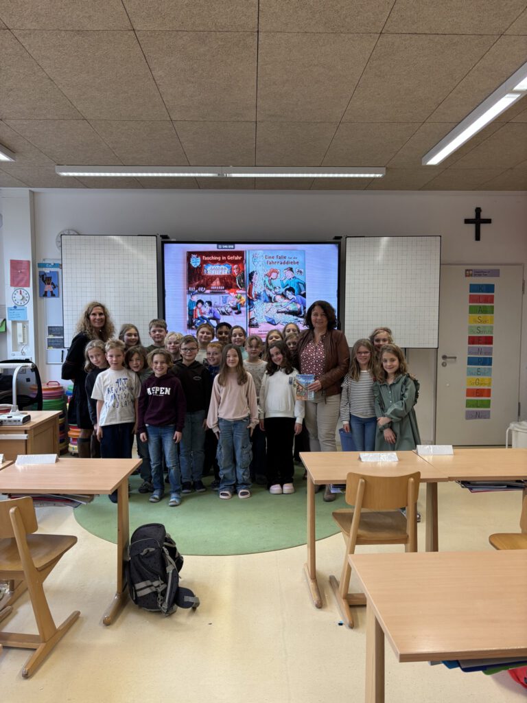 Frau Ruth Müller steht mir der Klasse 3b und der Klassleitung Frau Claudia Wimmer vor der Tafel, auf der das Buchcover der Isardetektive zu sehen ist