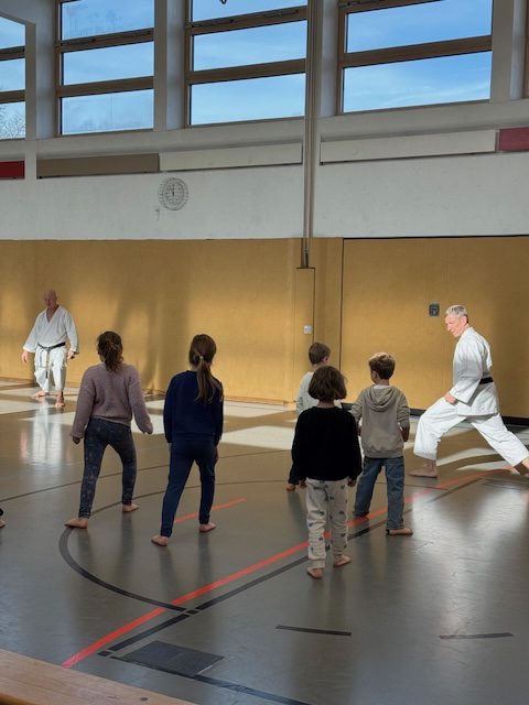 Kinder sind in der Turnhalle und führen mit zwei Karatetrainern Übungen durch