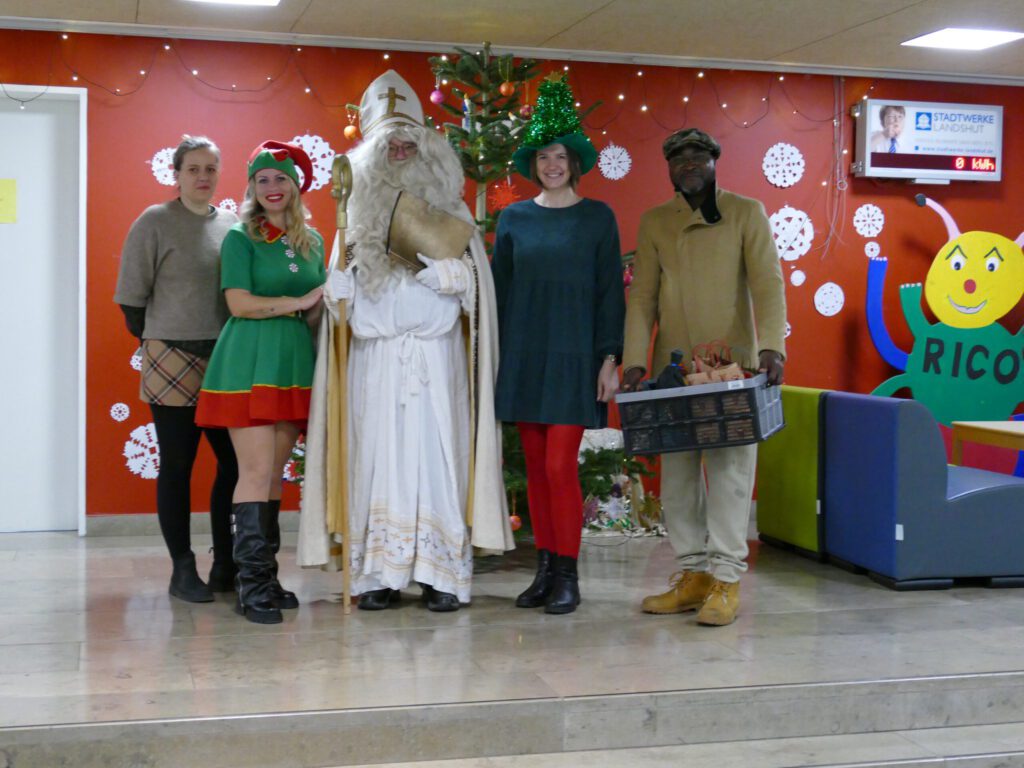 Der heilige Nikolaus steht mit zwei Weihnachtselfen und zwei weiteren Personen in der Aula der GS Carl-Orff