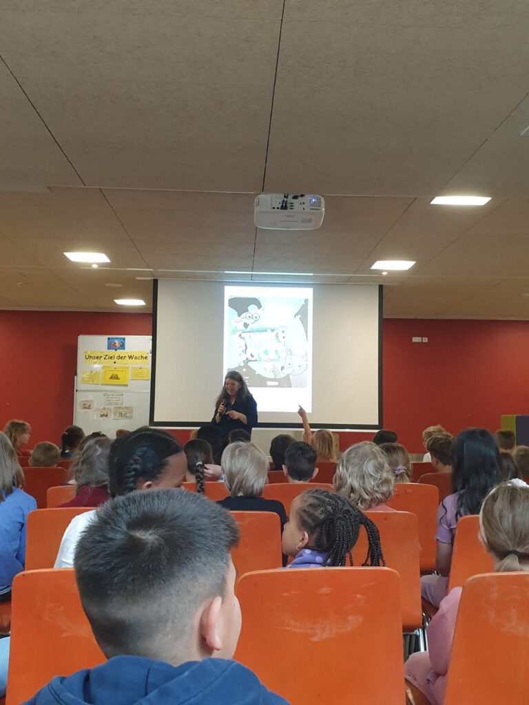 Kinder sitzen in der Aula, Autorin liest aus ihrem Werk vor