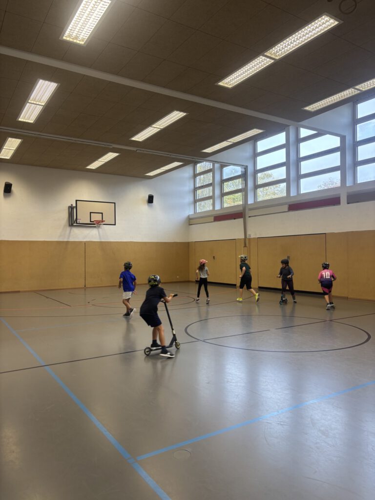 Kinder fahren Roller in einer Turnhalle, sie tragen Helme, manche Kinder laufen in der Halle