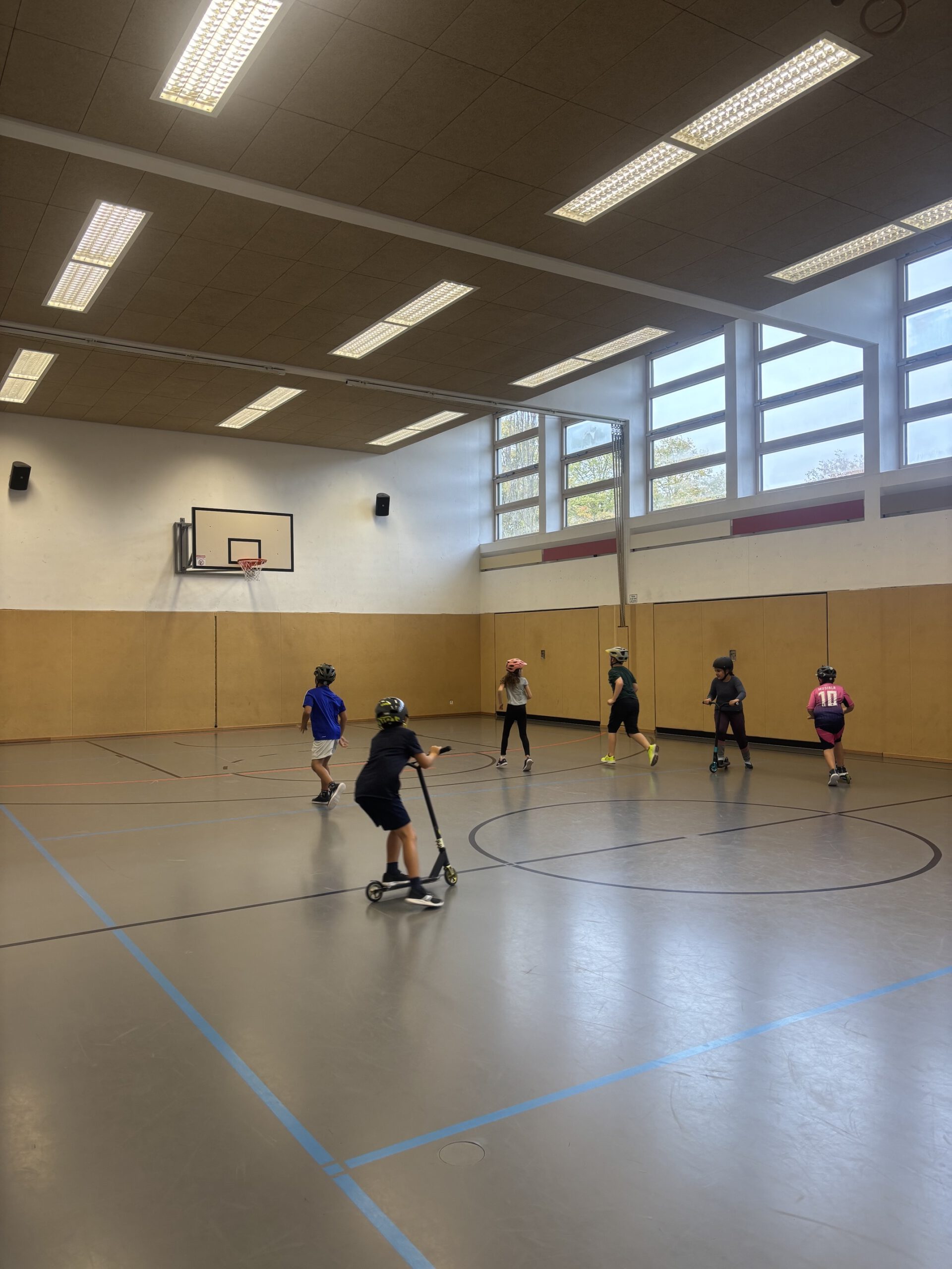 Kinder fahren Roller in einer Turnhalle, sie tragen Helme, manche Kinder laufen in der Halle